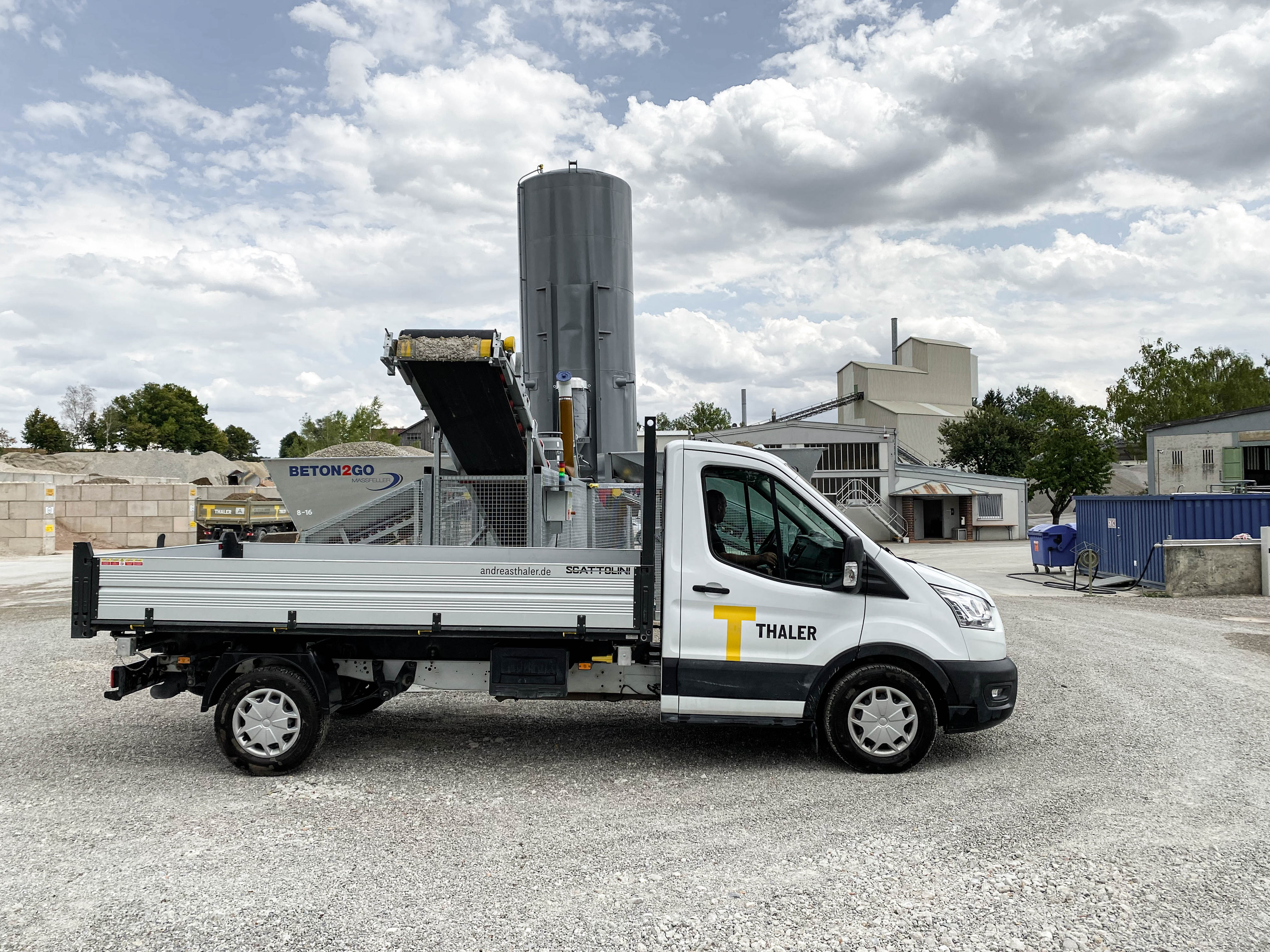 Ein weißer Thaler-Lkw parkt auf Schotter in einem Baustellengebiet mit Industriegebäuden und einem bewölkten Himmel im Hintergrund.