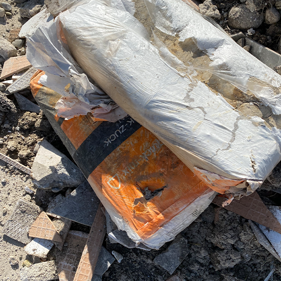 Ein beschädigter Zementsack liegt zwischen verstreutem Schutt und Trümmern im Freien.