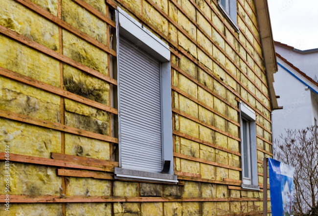 Nahaufnahme eines Hauses mit gelber Ziegelverkleidung und zwei kleinen Fenstern mit geschlossenen Fensterläden. Einige der Ziegel sind mit Moos bedeckt.