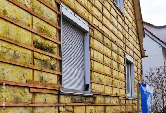 Nahaufnahme eines Hauses mit gelber Ziegelverkleidung und zwei kleinen Fenstern mit geschlossenen Fensterläden. Einige der Ziegel sind mit Moos bedeckt.