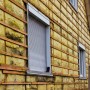 Nahaufnahme eines Hauses mit gelber Ziegelverkleidung und zwei kleinen Fenstern mit geschlossenen Fensterläden. Einige der Ziegel sind mit Moos bedeckt.