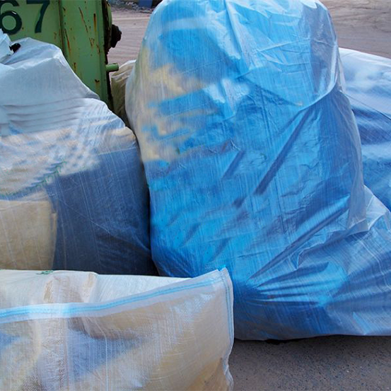 Große blaue und weiße Säcke mit wiederverwertbarem Material, gestapelt neben einem grünen Müllcontainer auf einer Betonfläche.