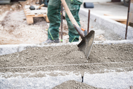 Nahaufnahme einer Person in grünen Arbeitshosen und -stiefeln, die mit einer Schaufel nassen Beton auf einer Baustelle glättet.