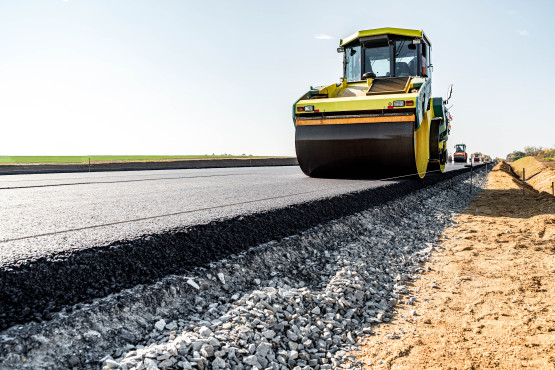 Eine Dampfwalze pflastert eine neue Asphaltstraße, mit einem klaren blauen Himmel und Kies am Straßenrand.