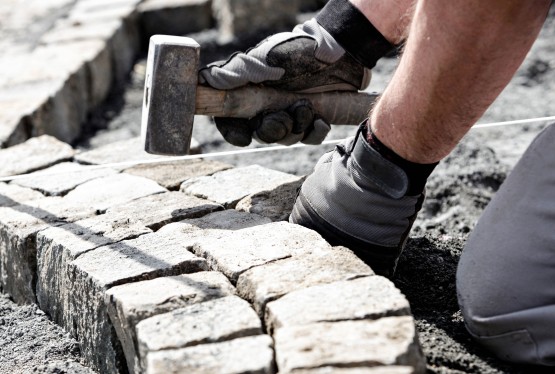 Ein Arbeiter verlegt Steinziegel in einem geschwungenen Muster. Er trägt Handschuhe und verwendet einen Hammer sowie eine Schnur zur Ausrichtung.