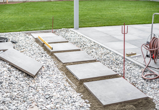 Betontrittsteine werden in einen Kiesweg neben einem Rasen verlegt. Eine Wasserwaage und Werkzeuge liegen in der Nähe.