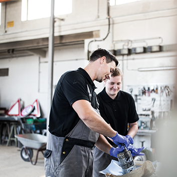 Zwei Mechaniker arbeiten in einer Werkstatt an einem mechanischen Teil, beide tragen schwarze Hemden und graue Overalls.