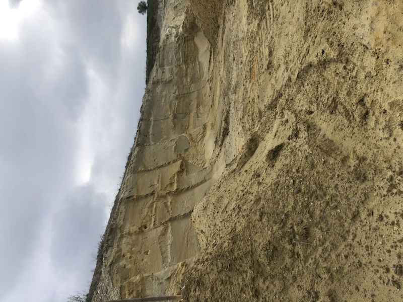 Erodierte Sandklippe unter einem bewölkten Himmel, darunter sind Schichten aus Sedimentgestein und losem Sand zu sehen.