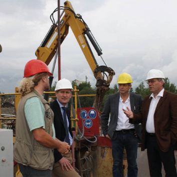 Vier Männer in Schutzhelmen und Geschäftskleidung unterhalten sich auf einer Baustelle mit Maschinen im Hintergrund.