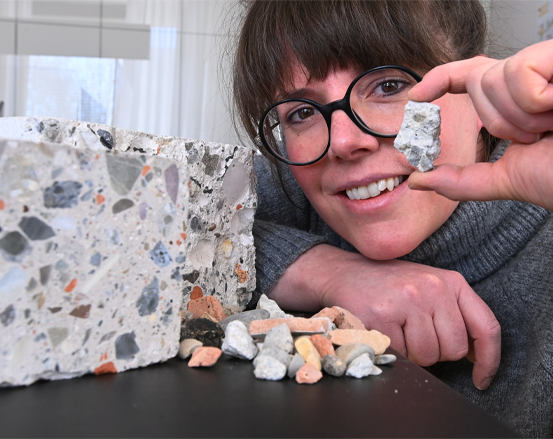 Eine Person mit Brille lächelt und hält ein kleines Stück Beton. Ein größerer Block und kleinere Stücke liegen auf einem Tisch.