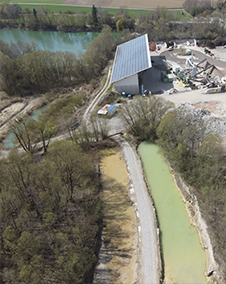 Luftaufnahme eines Industriegebiets am Flussufer mit einem großen Gebäude, umgeben von Grünflächen und Wasserkanälen.