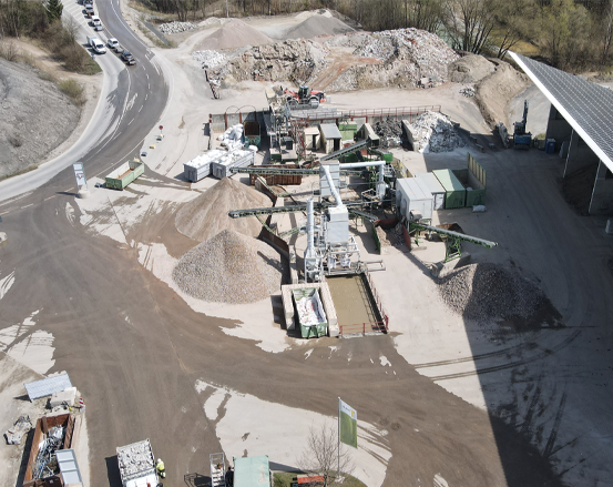 Luftaufnahme eines industriellen Recyclinggeländes mit Maschinen, Kieshaufen und Containern in der Nähe einer kurvenreichen Straße.