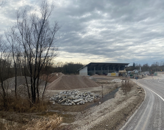 Baustelle mit Kies- und Schutthaufen in der Nähe eines teilweise fertiggestellten Gebäudes. Bewölkter Himmel und kahle Bäume im Hintergrund.