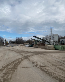 Industriegelände mit Maschinen, Kieshaufen und Förderband unter bewölktem Himmel. Bäume im Hintergrund.