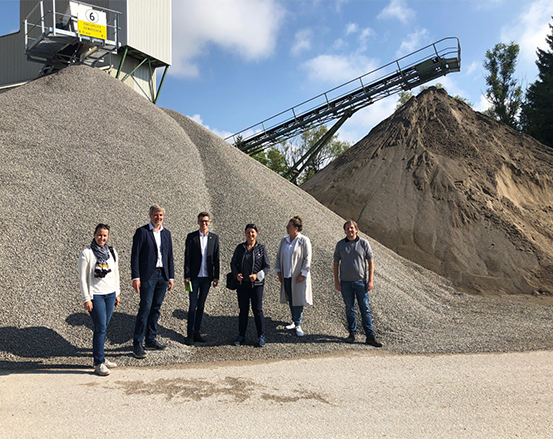 Eine Gruppe von Menschen steht vor großen Kies- und Sandhaufen auf einem Industriegelände im Freien.