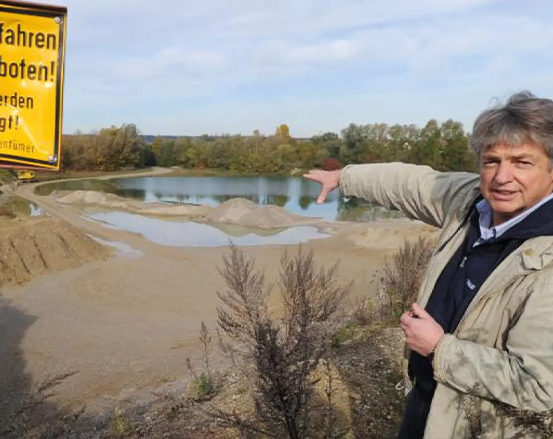 Eine Person deutet auf eine Kiesgrube und einen Teich. In der Nähe steht ein Warnschild auf Deutsch. Der Himmel ist teilweise bewölkt.
