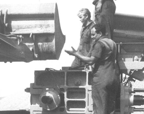 Drei Personen arbeiten im Freien an schweren Maschinen, wobei eine Person in Richtung der Maschine deutet.