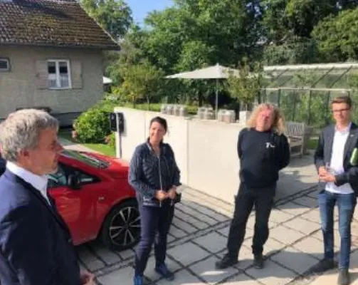 Eine Gruppe von vier Personen steht draußen neben einem roten Auto und unterhält sich in einem sonnenbeschienenen Gartenbereich.