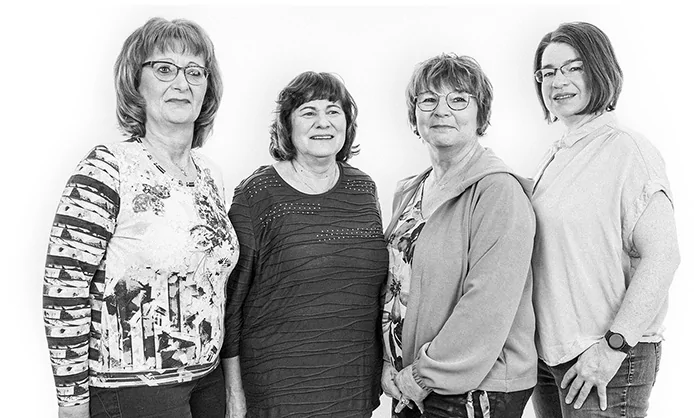 Vier lächelnde Frauen stehen nebeneinander in einem Gruppenporträt vor einem schlichten Hintergrund.