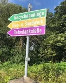 Wegweiser mit deutschem Text weisen auf Recycling, Kies-Sand-Werk und Betonstation vor einem grünen Hintergrund hin.